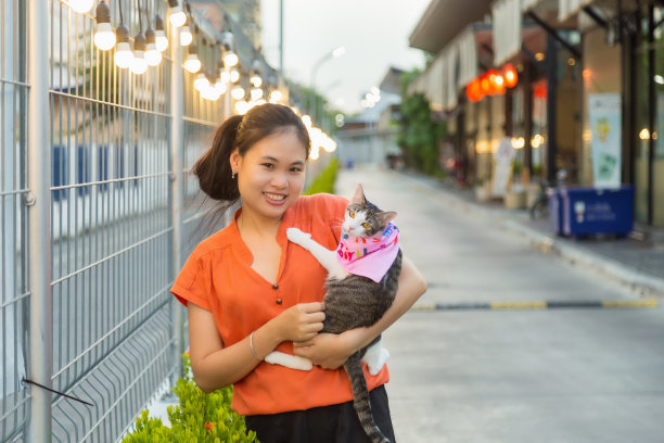 这是一幅微笑的年轻亚洲女子的肖像，她穿着橙色衬衫，抱着可爱的育种猫，她站在食物大道的金属墙旁边，在晚上。图片下载