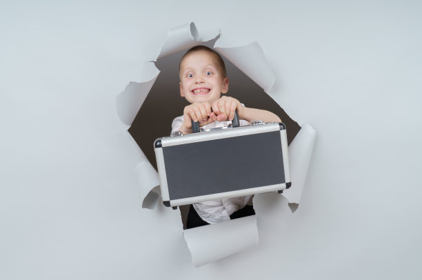 酷男孩提着一个装满钱的手提箱从工作室的破纸洞里冒了出来，带着兴奋愉悦的表情，透过灰色的背景看着。图片下载