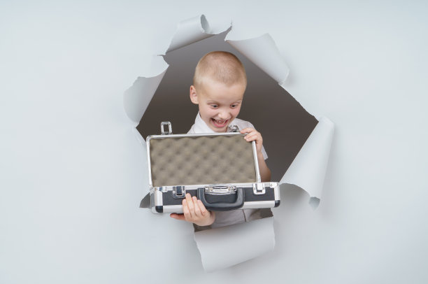 酷男孩提着一个装满钱的手提箱从工作室的破纸洞里冒了出来，带着兴奋愉悦的表情，透过灰色的背景看着。图片下载