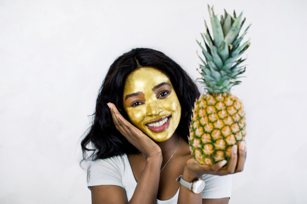 近距离美丽的非洲女人的肖像与金色的面具在脸上，拿着新鲜的菠萝和摆姿势在镜头与美好的微笑。护肤，清洁，有机化妆品图片下载