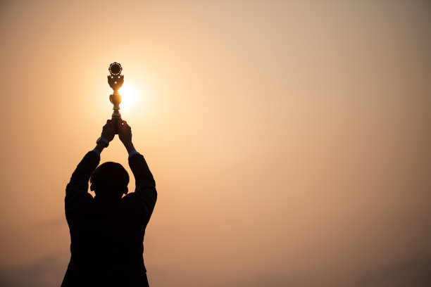 一个快乐的亚洲英俊的年轻商人在商业套装显示金灿灿的奖杯和持有文件夹在日落日出山在背景。经营理念。图片下载