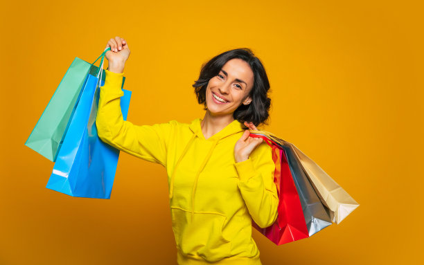 我终于得到了我想要的!一个年轻女子的半长照片，穿着黄色的帽衫，因为购物而高兴，拿着她在彩色的袋子里买的东西。图片下载