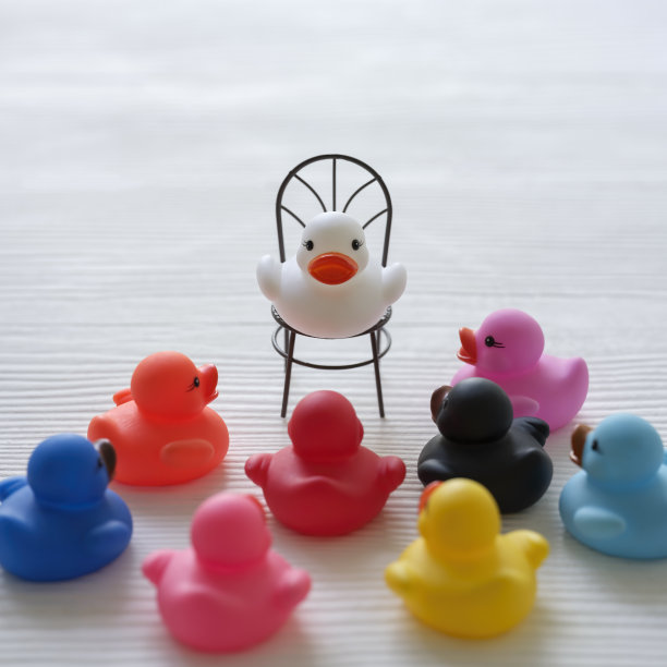一群五彩缤纷的橡皮鸭，围拢在一起，仿佛在听一只白色橡皮鸭坐在更高的椅子上，仿佛在布道，场景设置在白色木纹纹理的背景上。图片下载