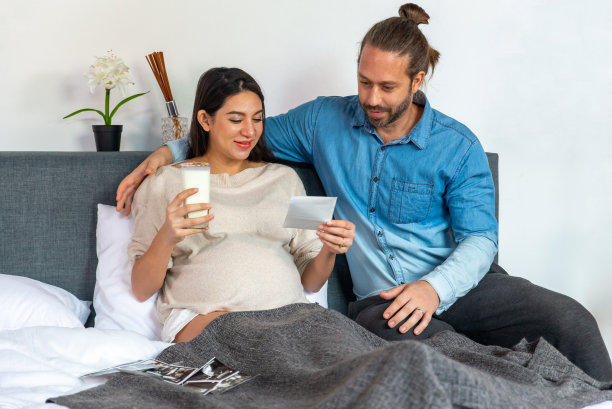 微笑年轻美丽的白种孕妇妻子喝新鲜牛奶和看着超声波婴儿照片在床上的丈夫在卧室。家庭纽带关系与孕产妇保健理念。图片下载