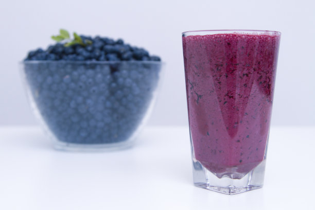 高脚杯与蓝莓冰沙的背景与一个大碗蓝莓在白墙上。图片下载