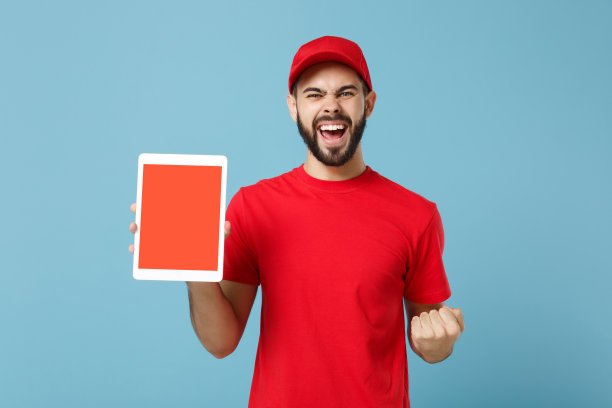 快递员身穿红色工作服，手持平板电脑隔离在蓝色背景，工作室肖像。专业男员工戴帽t恤工作快递经销商。服务的概念。模拟拷贝空间。图片下载