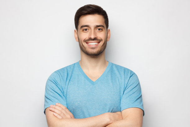 一个穿着蓝色t恤的年轻人，双臂交叉站在那里，微笑着，充满自信，孤立在灰色的背景下图片下载