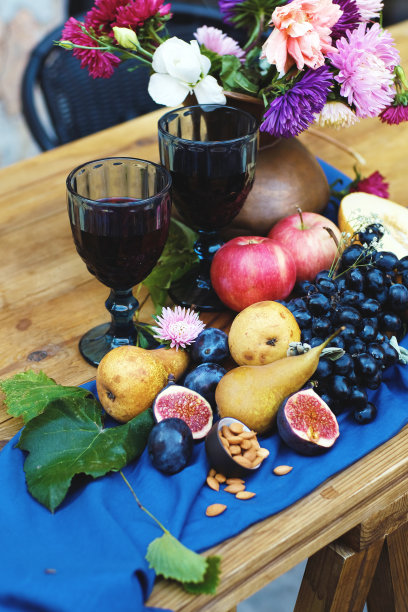蓝台布上有一幅静物画，里面有不同的水果和花瓶里的一束花。酒杯，葡萄，苹果，梨，无花果和紫花图片下载