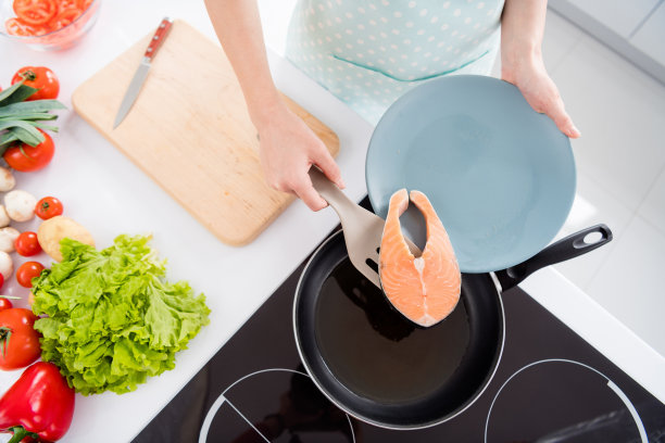 主妇夫人的裁剪照片，把新鲜生鲑鱼鱼片，翻过来躺着煎锅，烹饪家庭美味的晚餐，穿烹饪围裙，站在现代厨房室内图片下载