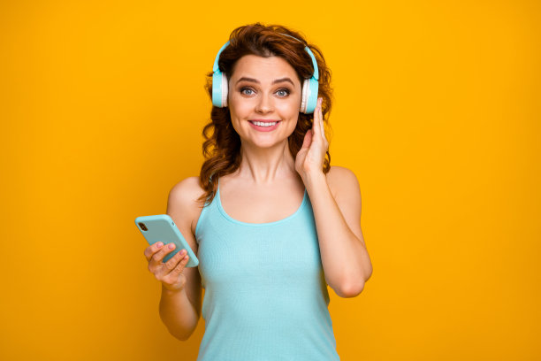 漂亮有趣的女士的照片拿电话听现代科技耳机流行歌曲好心情穿休闲的蓝绿色背心孤立的充满活力的黄色背景图片下载