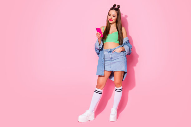 完整的照片漂亮的女士拿着电话发短信的男朋友穿牛仔夹克绿色的露脐装裸露的肚子牛仔衬衫膝盖袜子鞋子孤立的粉颜色的背景图片下载