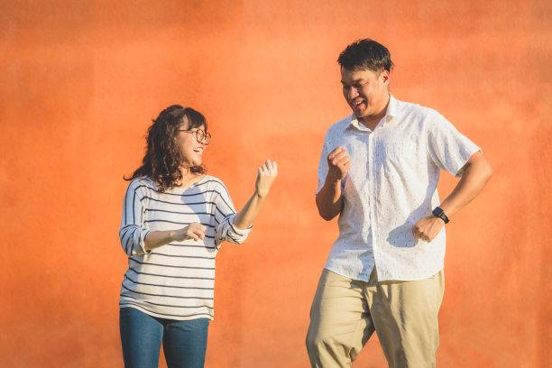 快乐的年轻亚洲夫妇的肖像一起大笑和跳舞在橙色的墙壁背景，夫妇放松和花时间在一起，人和生活理念图片下载