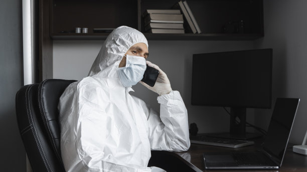 在隔离期间，身着防护服、佩戴医用口罩和橡胶手套的男子坐在家里与朋友或商业伙伴通电话。在新型冠状病毒流行中远程工作的人。冠状病毒图片下载