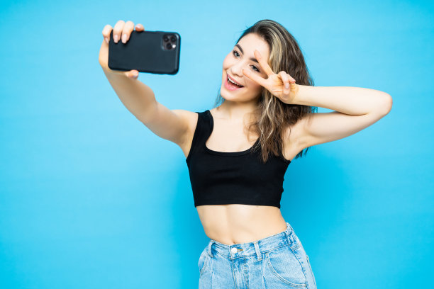一个年轻有魅力的女人的肖像与和平手势使自拍照片与智能手机隔离在蓝色背景图片下载