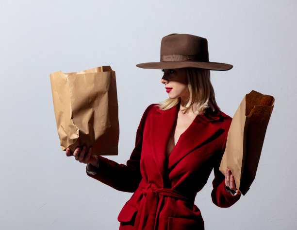 金发女孩穿着红色外套，戴着复古帽子，灰色背景上有超市纸袋。图片下载