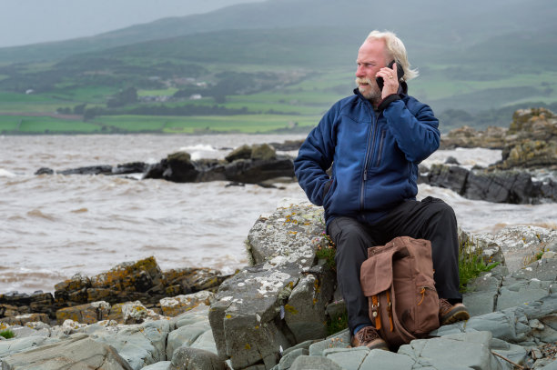 一个多风的阴天，一个活跃的老人坐在海边用手机图片下载
