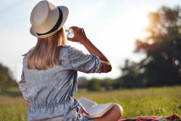 戴着帽子的女人坐在毯子上喝着瓶装水，在日落的大自然中。放松和休息的概念图片下载