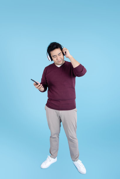 快乐的亚洲男人穿着毛衣，用耳机和智能手机在蓝色背景。图片下载
