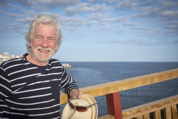 有魅力的快乐老人戴着草帽和蓝色条纹衬衫在海上远足，靠在木栈道的栏杆，蓝天和云彩的背景-积极退休老人的概念图片下载