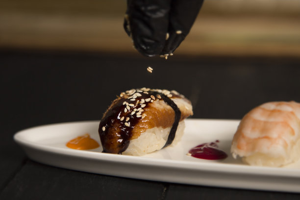 日本手握寿司的芝麻配鳗鱼，米饭和酱汁放在白色盘子里。厨师手戴防护手套。图片下载