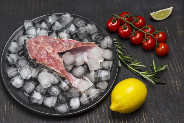 在黑色陶瓷盘子上融化的冰块上的新鲜河鱼。柠檬，樱桃番茄和迷迭香。图片下载