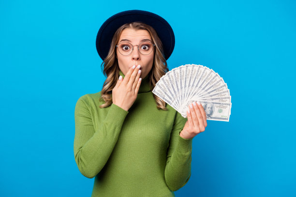 疯狂滑稽的卷发女士的照片持有风扇美元美元富人自由职业者的薪水隐藏嘴手臂面部表情戴眼镜帽子绿色高领孤立的蓝色背景图片下载