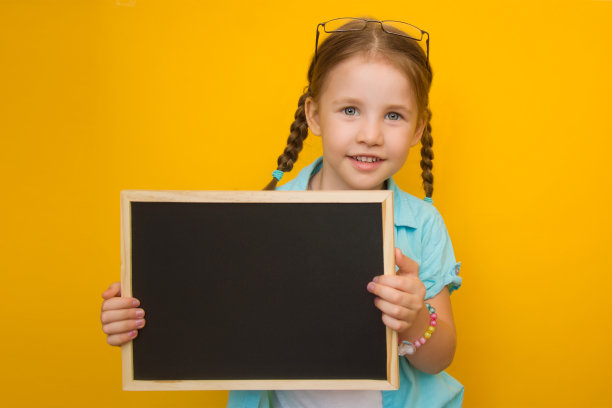 回到学校。漂亮的小女孩扎着辫子，戴着眼镜，手里拿着一块黄色背景的黑板。Copyspace图片下载