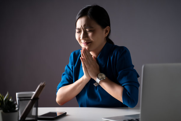 亚洲女人幸福在蓝色衬衫牵手祈祷，看着复印空间，工作在一个笔记本电脑在办公室。图片下载