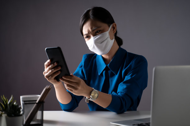 亚洲女性戴着防护面具看智能手机看新闻，在办公室感到困惑。孤立的背景。图片下载