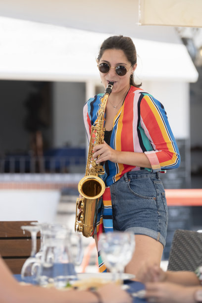 一个戴着眼镜的年轻女子，在一个模糊的户外酒吧露台旁吹着萨克斯风图片下载