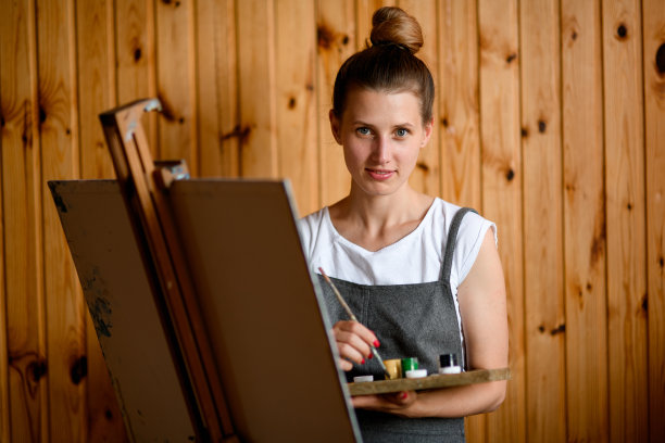 美丽的年轻女子，手拿调色板和画笔，站在画架旁边看着相机。图片下载