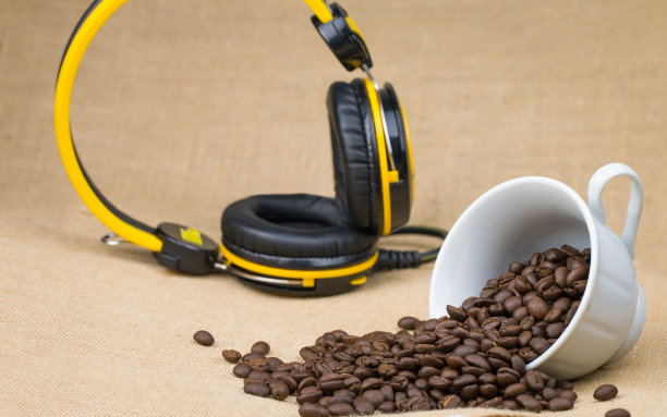 在一个白色的咖啡杯在它的一边和许多咖啡豆散落在地板上和音乐耳机放在后面的图片和底部表面是一个棕色的织物。图片下载