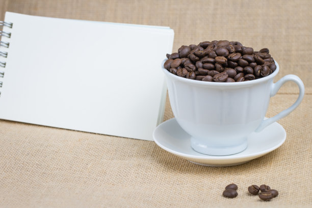 在一个白色的咖啡杯和一个空白的笔记本，底面是一个棕色的织物和一些咖啡豆散落在地板上，图片有一个空白的空间图片下载