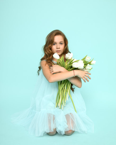 一个美丽的女孩卷着头发跪在蓝色的背景上，拿着白色的郁金香的工作室照片。节日的问候。妇女节。老师的一天。感激之情。自然美景。柔软干净的皮肤图片下载