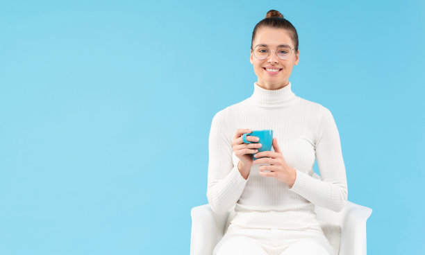 年轻的女孩戴着眼镜，手里拿着一杯茶，坐在白色的扶手椅上休息，在蓝色的背景上孤立图片下载