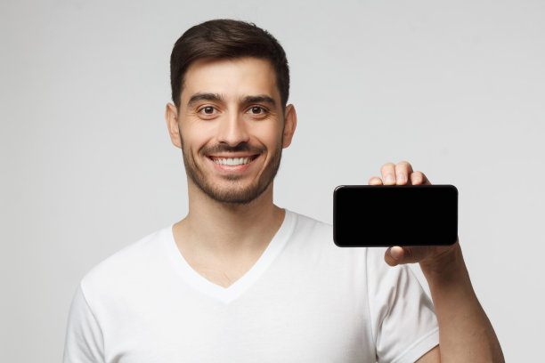 特写肖像的年轻人穿着白色t恤拿着空白屏幕手机在水平位置，显示给观众，孤立在灰色背景图片下载