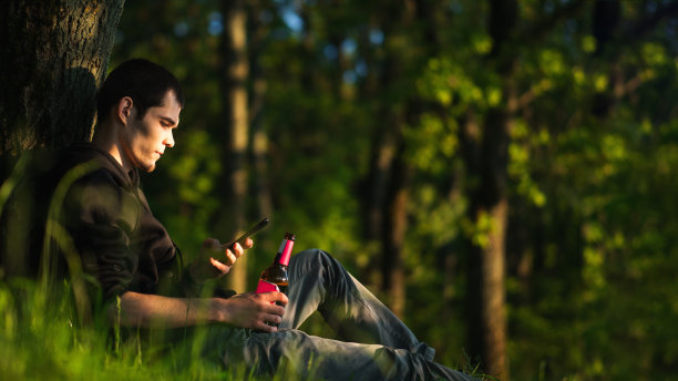 一个年轻人倚靠在公园的一棵树上躺在草地上，手里拿着一瓶啤酒图片下载