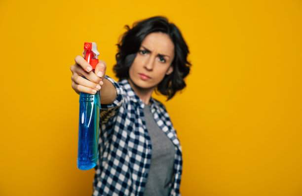 呆在原地别动。一个严格的漂亮女孩的特写照片，在一个格子衬衫，指着一个蓝色瓶子喷在相机上，看着威胁。图片下载