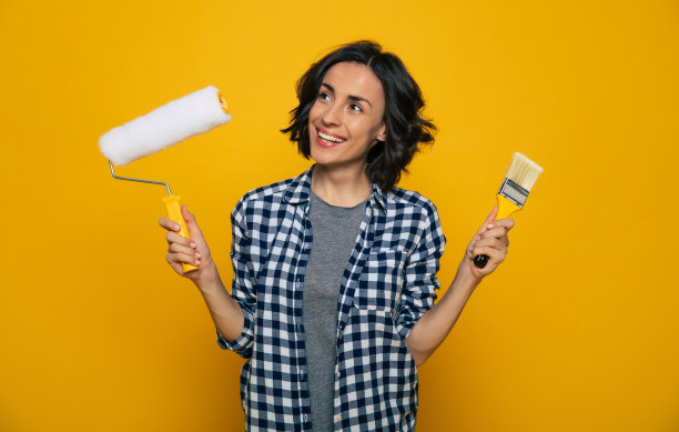 绘画的一天。一张漂亮女孩的半长照片，穿着格子衬衫，右手拿着油漆滚筒，左手拿着刷子，眼睛明亮地望着一边。图片下载