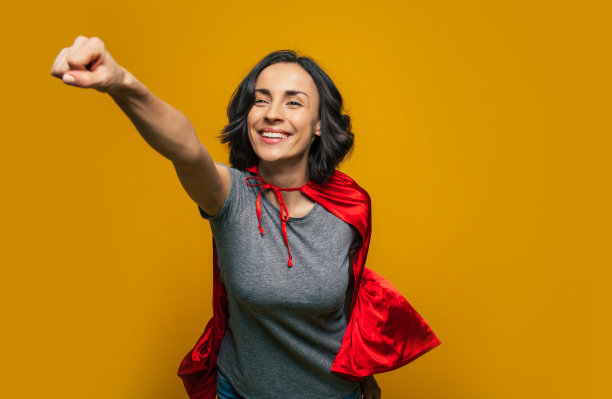 我在路上了!一个年轻美丽的女孩，假装自己是一个女超人，并在一个经典的超人姿势飞行，穿着休闲，但与超人红色斗篷。图片下载