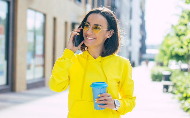 有什么新鲜事吗?这是一个可爱的女孩的特写照片，她穿着黄色的连帽衫，戴着眼镜，手里拿着白色的智能手表，用她的现代手机说话。图片下载