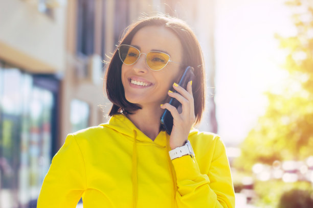 让我们谈谈。这是一个穿着黄色连帽衫、戴着眼镜的时尚微笑女孩的特写照片，她手里拿着智能手表，一边看一边用她的时髦手机说话。图片下载