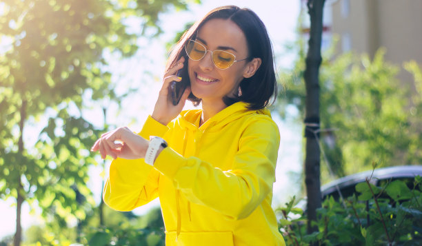 那我们什么时候见面?这是一个现代都市女孩的特写照片，她穿着黄色的连帽衫，戴着眼镜，涂着黄色的指甲，一边打电话一边看智能手表。图片下载