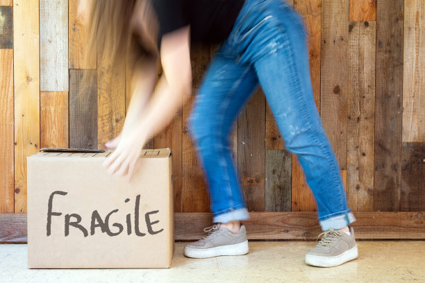 年轻女子带着搬家箱走在新家靠近木墙的地方，在搬家的日子里拿着硬纸板上写着易碎的复古设计搬家箱图片下载
