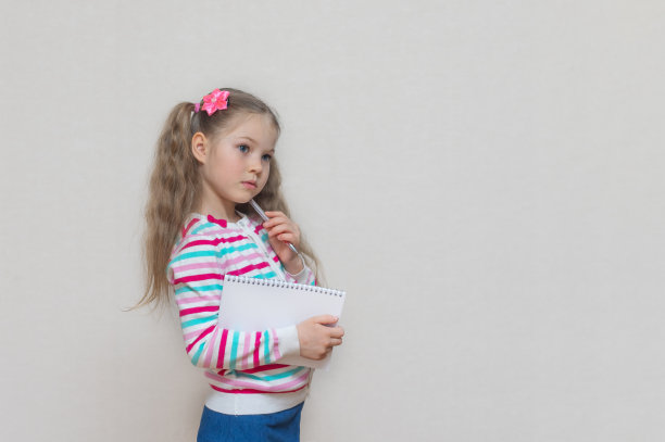 回到学校。一个金发女学生的肖像，手里拿着笔记本和笔。她考虑了这个任务。浅灰色工作室背景。教育。复制的地方。图片下载