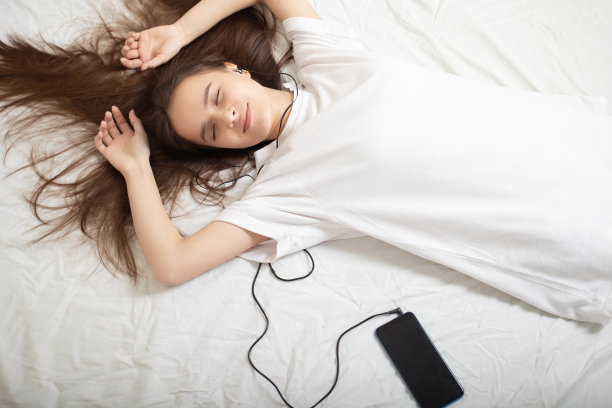 音乐的生活方式。女孩戴着耳机，闭着眼睛看手机躺在床上图片下载