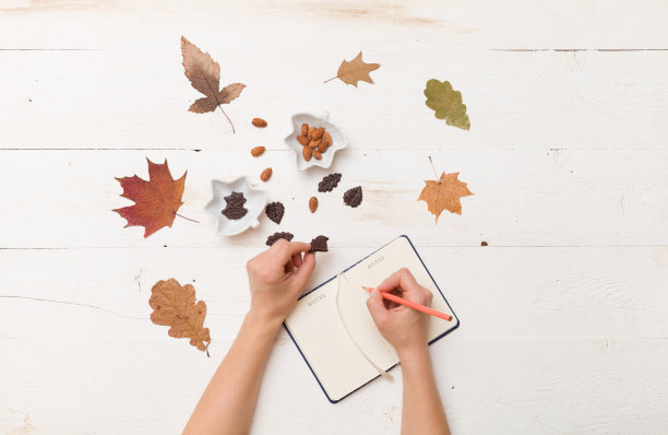 俯视图女人的手写在笔记本上。秋天的背景:树叶，巧克力，坚果木制白板。图片下载