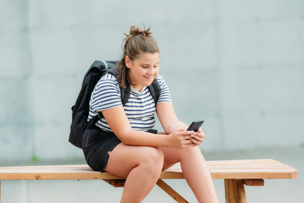 一个面带微笑的13岁学生坐在她学校前面的一张图片桌子上写短信。图片下载
