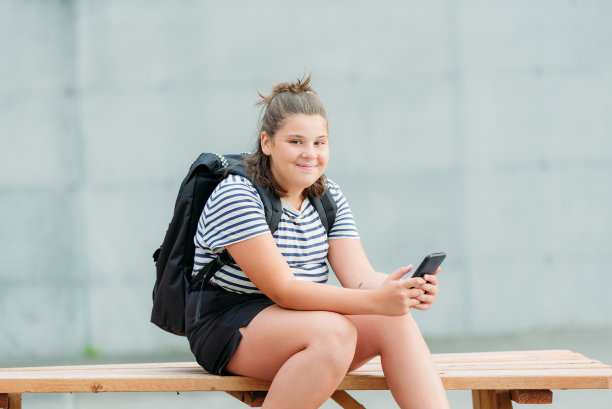 一个面带微笑的13岁学生坐在她学校前面的一张图片桌子上写短信。图片下载