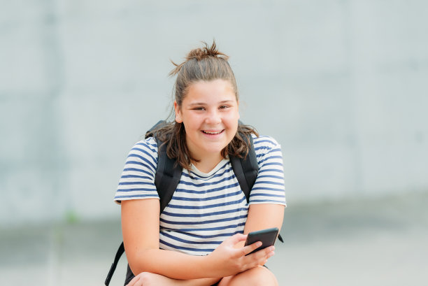 一个笑着的13岁的学生坐在她学校前面的一张图片桌子上写短信。图片下载